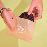 Vegan Raspberry Dark Choc New York Cookie being held in Crumbs & Doilies branded packaging against a light background